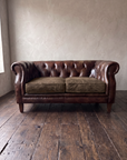 Vintage tobacco leather chesterfield loveseat, front view, greige plaster wall and dark reclaimed wood floor