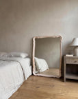 Blush serpentine mirror leaning against a plaster bedroom wall between a linen-made bed and a wooden nightstand