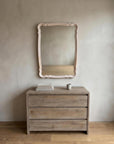 Blush French Provincial style mirror hung above a wooden dresser with books and a ceramic cup