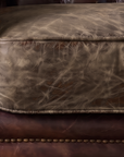 Macro of the smoky brown leather seat cushion with pale silvery crackle, welt and brass nailhead rail below