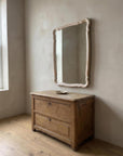 Blush serpentine mirror hung above an antique wooden chest beside a window, seen at an angle
