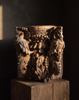 Carved teak temple capital on a wood plinth in a dark gallery with a shaft of warm light