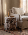 Carved teak temple capital used as a low side table beside a linen chair with a candle and book