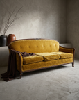 Vintage carved-wood camelback sofa in gold crushed velvet, styled in a greige plaster room with dried branches and draped chocolate linen