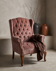 Dusty rose velvet tufted wingback chair, lifestyle view in a plaster-walled room with stoneware vessels and dried branches