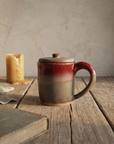 Lidded stoneware mug with deep red glaze breaking into warm sand, round loop handle, on a weathered oak table