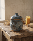 Blue-grey lidded stoneware jar with raised scrollwork and unglazed clay bands, on a weathered oak table in warm afternoon light
