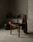 Sundays walnut dining chair with black leather seat, staged in a greige plaster room with stoneware vessels and dried branches