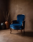 The Blue Velvet Bergere, deep sapphire velvet French chair with carved wood frame, in a rich brown plaster room