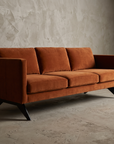 Burnt orange velvet boomerang-leg sofa, 45 degree three-quarter angle showing the track-arm form