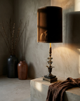 The Vesper Lamp lit on a greige plaster ledge, chocolate velvet drum shade glowing, stoneware vessels and bare branches beside it