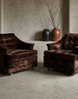 A pair of vintage chocolate crushed velvet club chairs angled toward each other in a greige troweled plaster room, with charcoal and terracotta stoneware vessels and bare branches behind