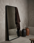 Large leaning floor mirror with matte black frame in a greige plaster room, styled with stoneware vessels and draped linen