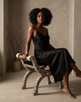 Woman with natural curls in a black silk slip dress seated on the bone inlay curule bench, showing the bench's true petite scale