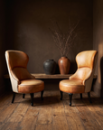 Pair of cognac leather and linen wingback slipper chairs in a dark chocolate plaster room with reclaimed wood floor