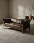 Cognac leather track-arm sofa, worn and lived-in patina, three-quarter lifestyle view in a greige plaster room with stoneware vessels
