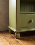 Close-up of a drawer with a round painted knob and a reeded bun foot on a wood floor