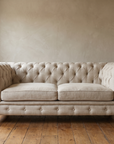 Natural oat Belgian linen Chesterfield sofa, straight-on front view in a plaster-walled room with a reclaimed wood floor