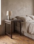 Vintage Indian side table in dark reclaimed wood used as a bedside table beside a linen-dressed bed, ceramic lamp and books on top, plaster wall and reclaimed wood floor