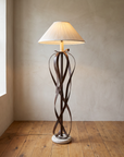 Ribbon floor lamp in hammered steel with a pleated shade, lit, against a plaster wall on a reclaimed wood floor