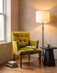 Forged iron tripod floor lamp switched on, glowing over a chartreuse velvet lounge chair, with a blackened wood side table and a steaming cup of tea