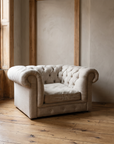 The chesterfield chair-and-a-half in the corner of a quiet prewar apartment room beside a tall window