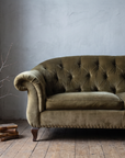 Olive velvet tufted sofa in a cool grey plaster corner with a bare branch and old books