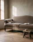 Taupe camelback sofa in soft morning window light with a linen throw over the arm and coffee on a wooden stool