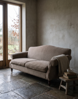 Taupe camelback sofa in a late autumn room with grey-brown plaster, stone floor and a bare garden beyond the window