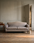 Taupe camelback sofa in a Parisian boiserie salon with herringbone parquet and doors open to amber light