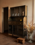 Steel hutch in a warm golden-lit room with blue and white plates behind the glass, an oak stool and a stone urn of dried grasses