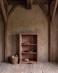 The bookcase in a tall barn holding a carved wooden bowl, old leather books and a stone, beside a basket of logs.