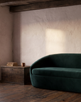 Long green velvet curved sofa in an old farmhouse room with a shuttered window, beams and a wooden chest