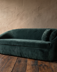 Deep green velvet curved tub sofa at a three-quarter angle against a rough taupe plaster wall on dark reclaimed wood floor
