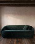 Front view of a green velvet curved sofa under dark timber beams with a small wooden stool and clay cup