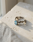 Sterling silver twig ring set with an oval blue topaz, laid flat on pale stone