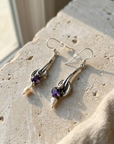 Sterling silver drop earrings with amethyst and baroque pearls, laid flat on pale stone