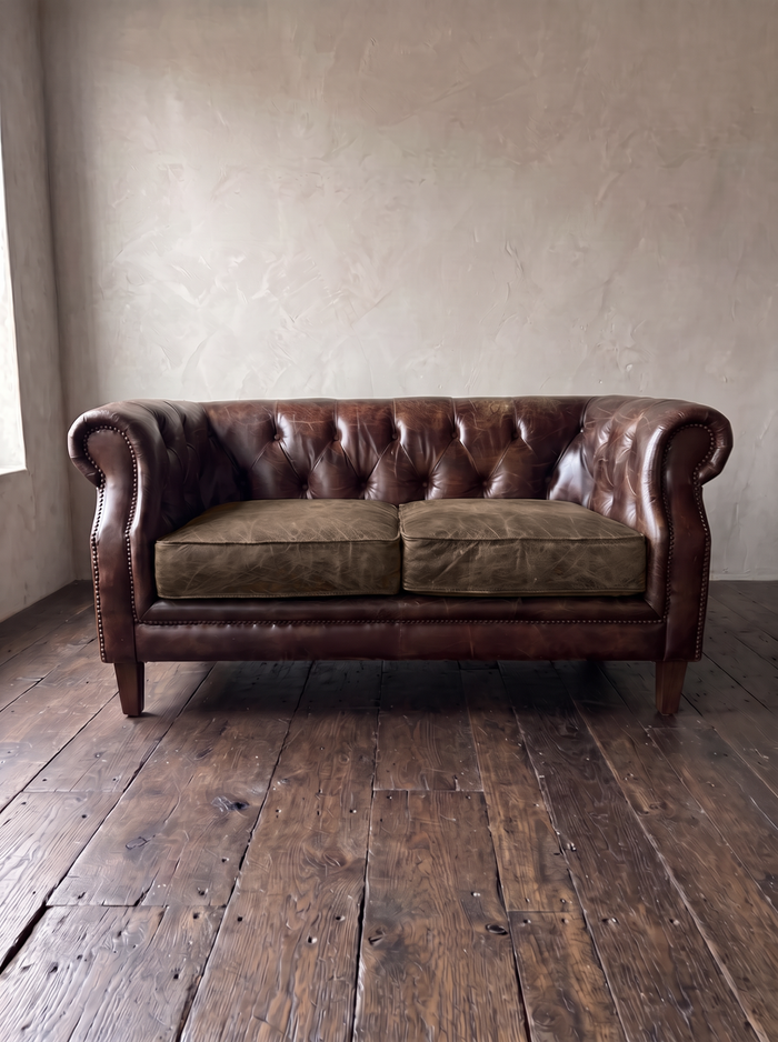 Vintage tobacco leather chesterfield loveseat, front view, greige plaster wall and dark reclaimed wood floor