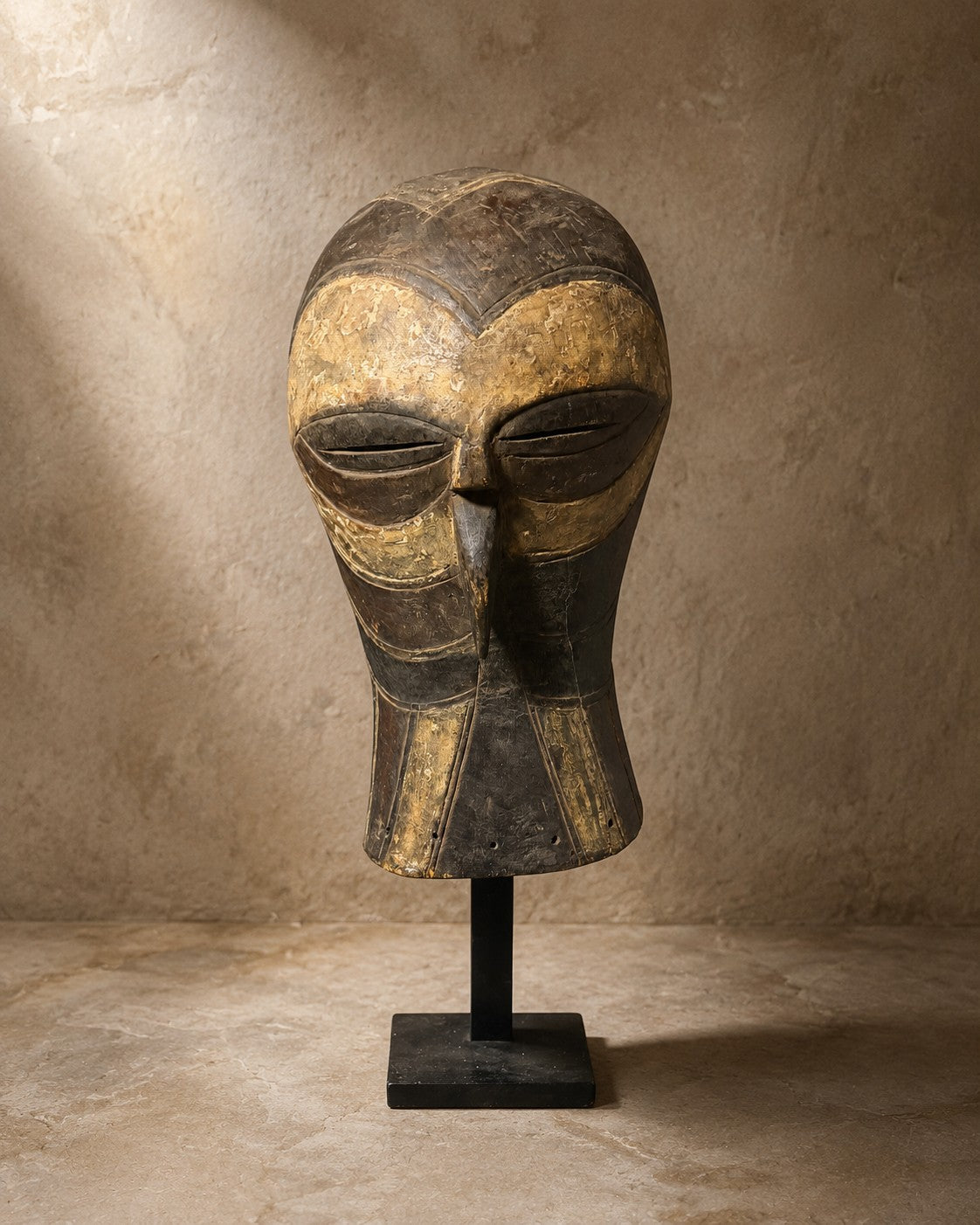Carved wooden mask with ochre pigment on a black display stand against a plaster wall
