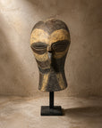 Carved wooden mask with ochre pigment on a black display stand against a plaster wall