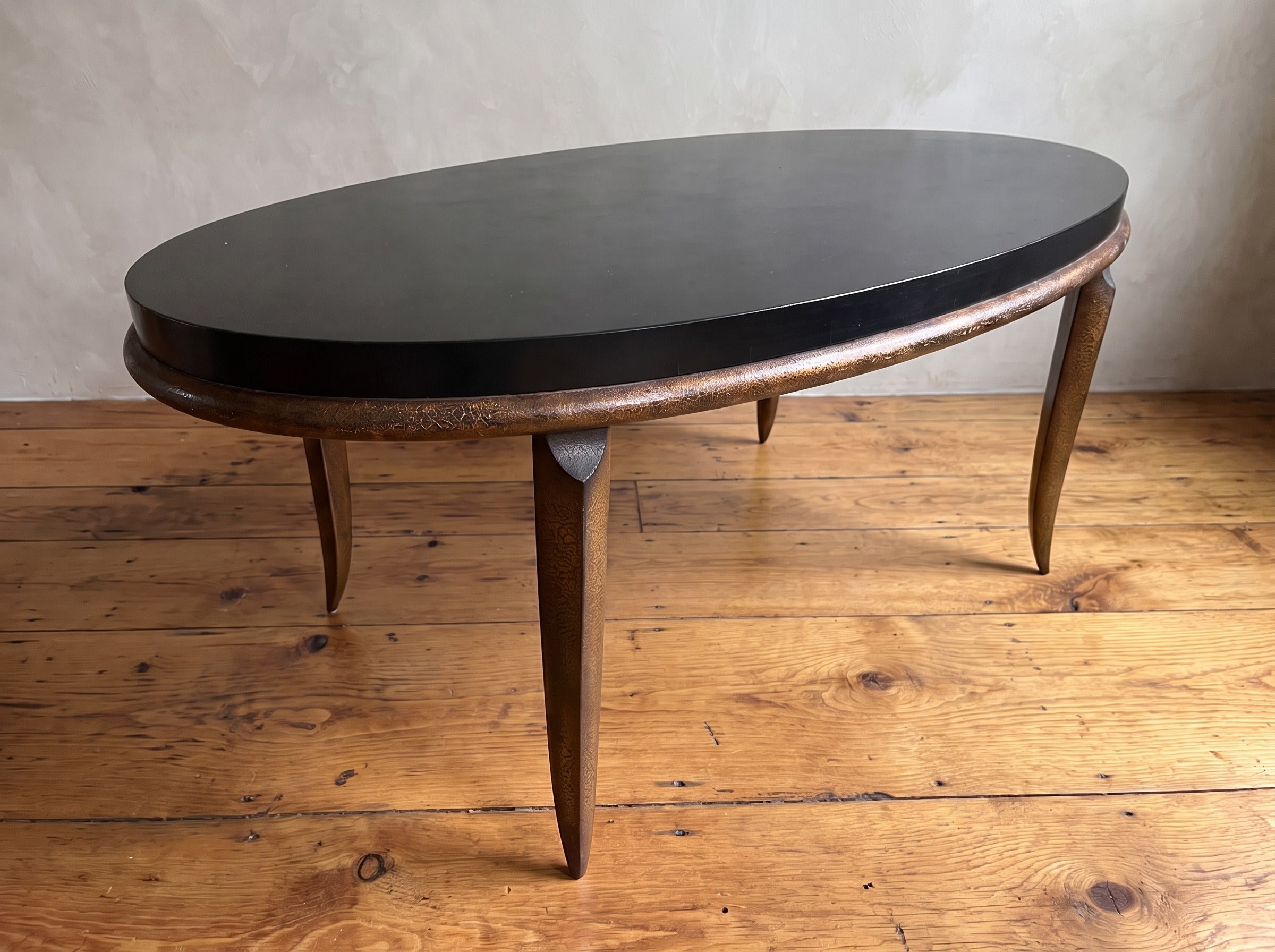 Vintage black and bronze oval coffee table with saber legs, three-quarter view, in a plaster-walled room on a reclaimed wood floor