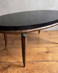 Vintage black and bronze oval coffee table with saber legs, three-quarter view, in a plaster-walled room on a reclaimed wood floor