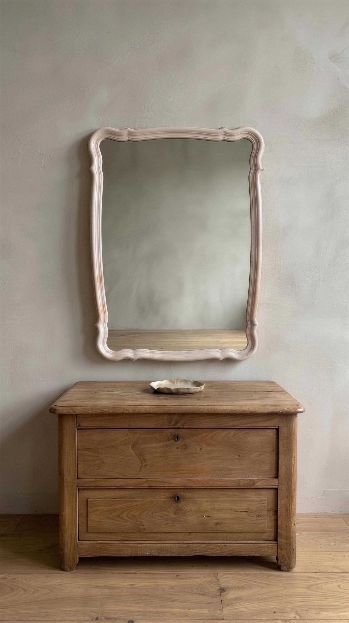 Blush French Provincial style mirror hung on a greige plaster wall above an antique wooden chest with a small ceramic dish