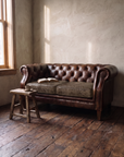 Leather chesterfield loveseat in morning window light with an open book and a stool with coffee