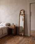 Gilt scroll mirror leaning beside a walnut nightstand and linen bed in a plaster bedroom