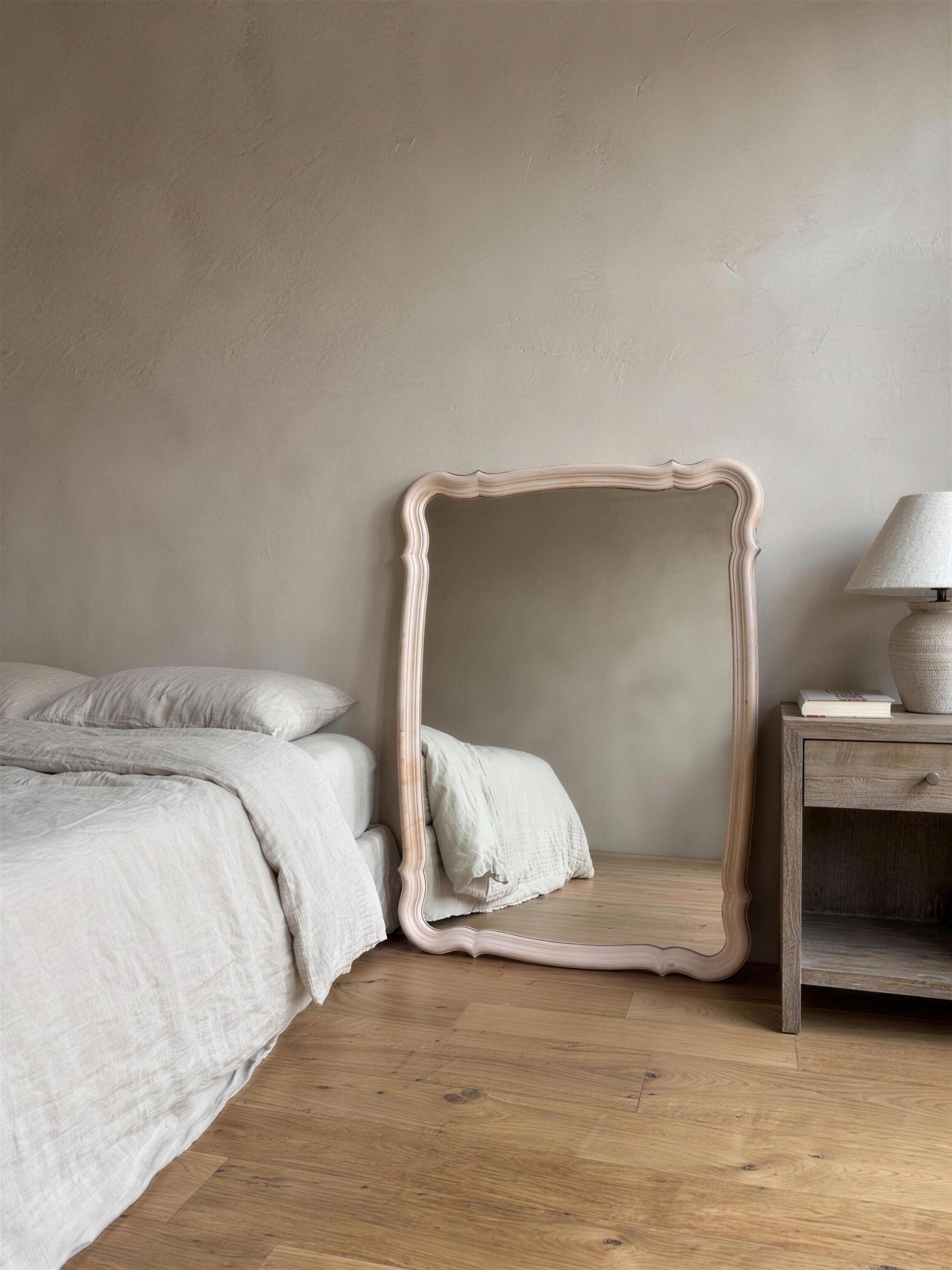 Blush serpentine mirror leaning against a plaster bedroom wall between a linen-made bed and a wooden nightstand