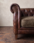 Close-up of the scroll arm, antique brass nailheads, crackled smoky brown leather cushion and tapered leg