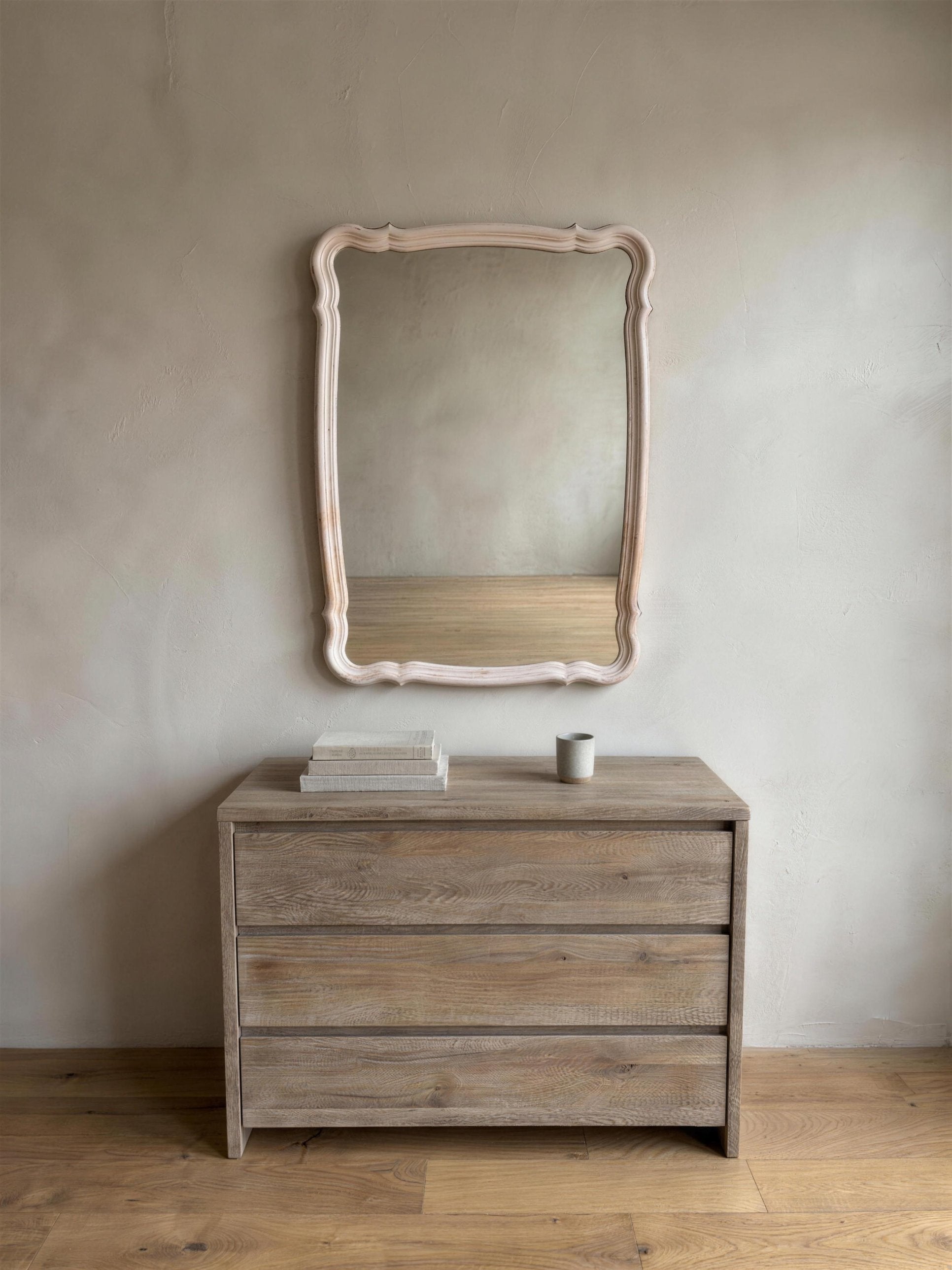 Blush French Provincial style mirror hung above a wooden dresser with books and a ceramic cup