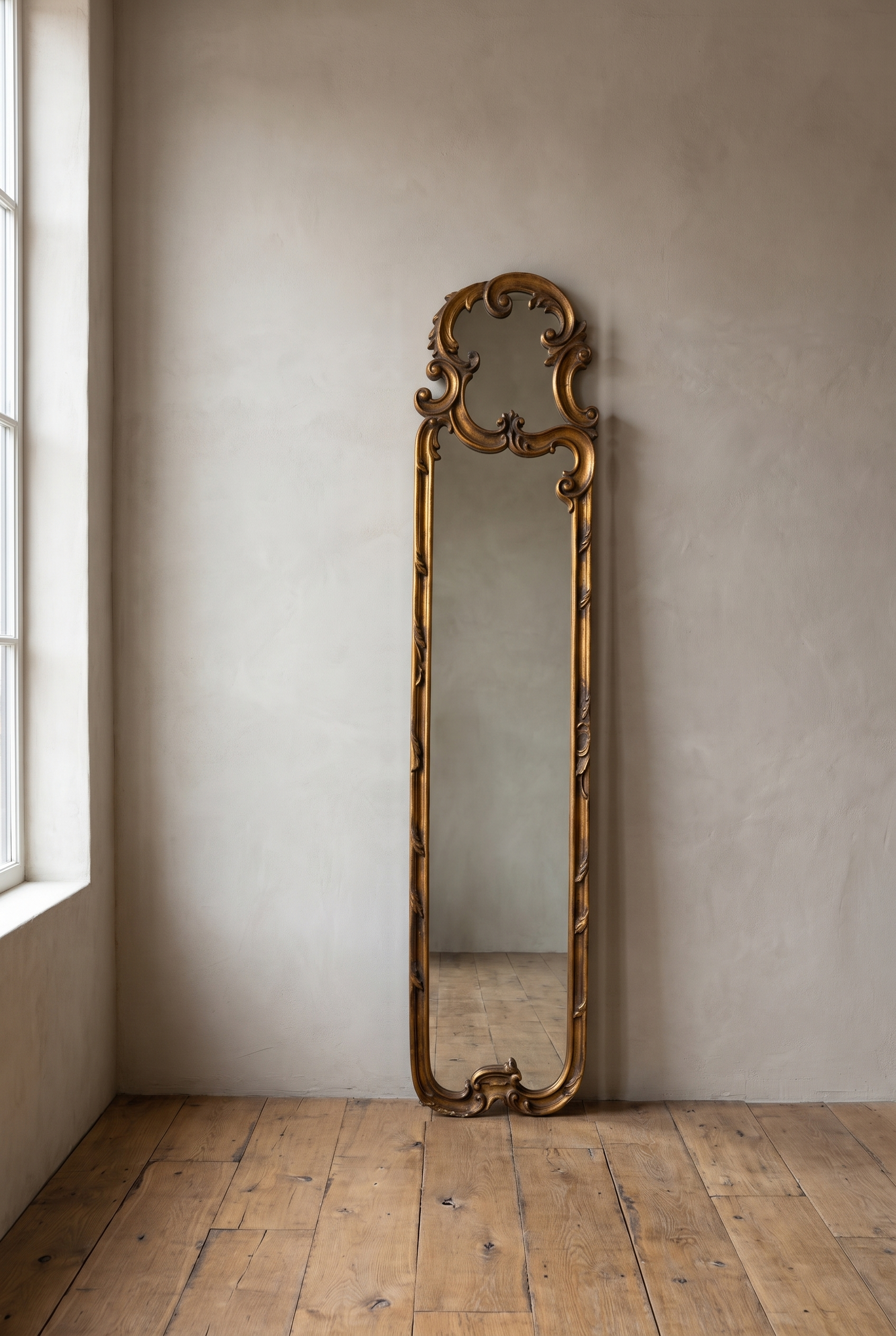 Tall narrow gilt rococo scroll mirror leaning against a warm plaster wall on a reclaimed wood floor
