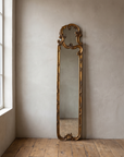 Tall narrow gilt rococo scroll mirror leaning against a warm plaster wall on a reclaimed wood floor